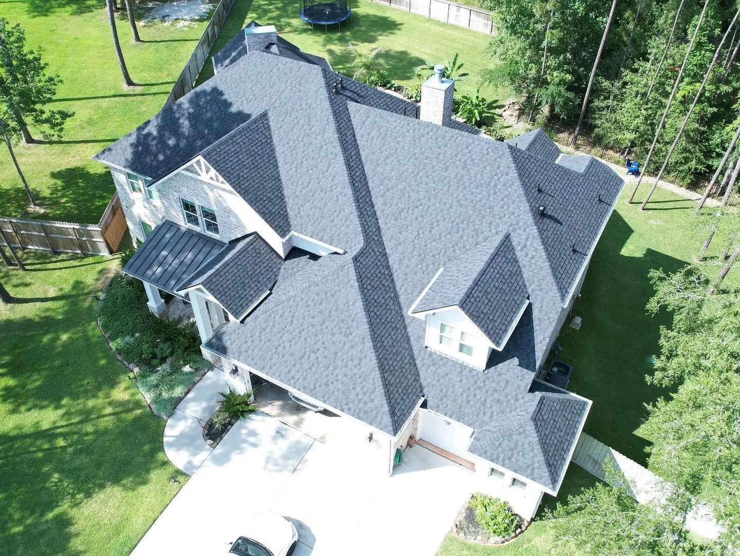 Aerial view of a dark gray shingled roof on a two-story house with white siding and lush green yard.