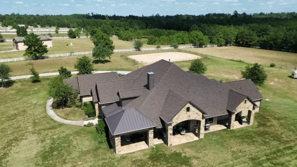 Large brick house with brown roof on grassy land, surrounded by trees and a rural landscape.