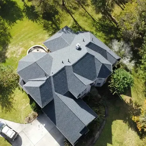 Overhead view of a house with a complex, dark gray roof, surrounded by green trees and grass.
