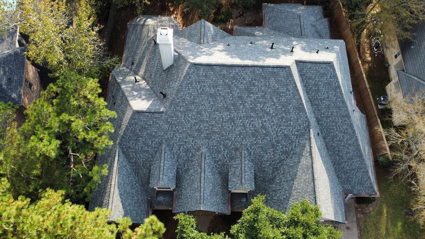 Aerial view of a house with a gray shingle roof, three dormers, and a white chimney, surrounded by green trees.