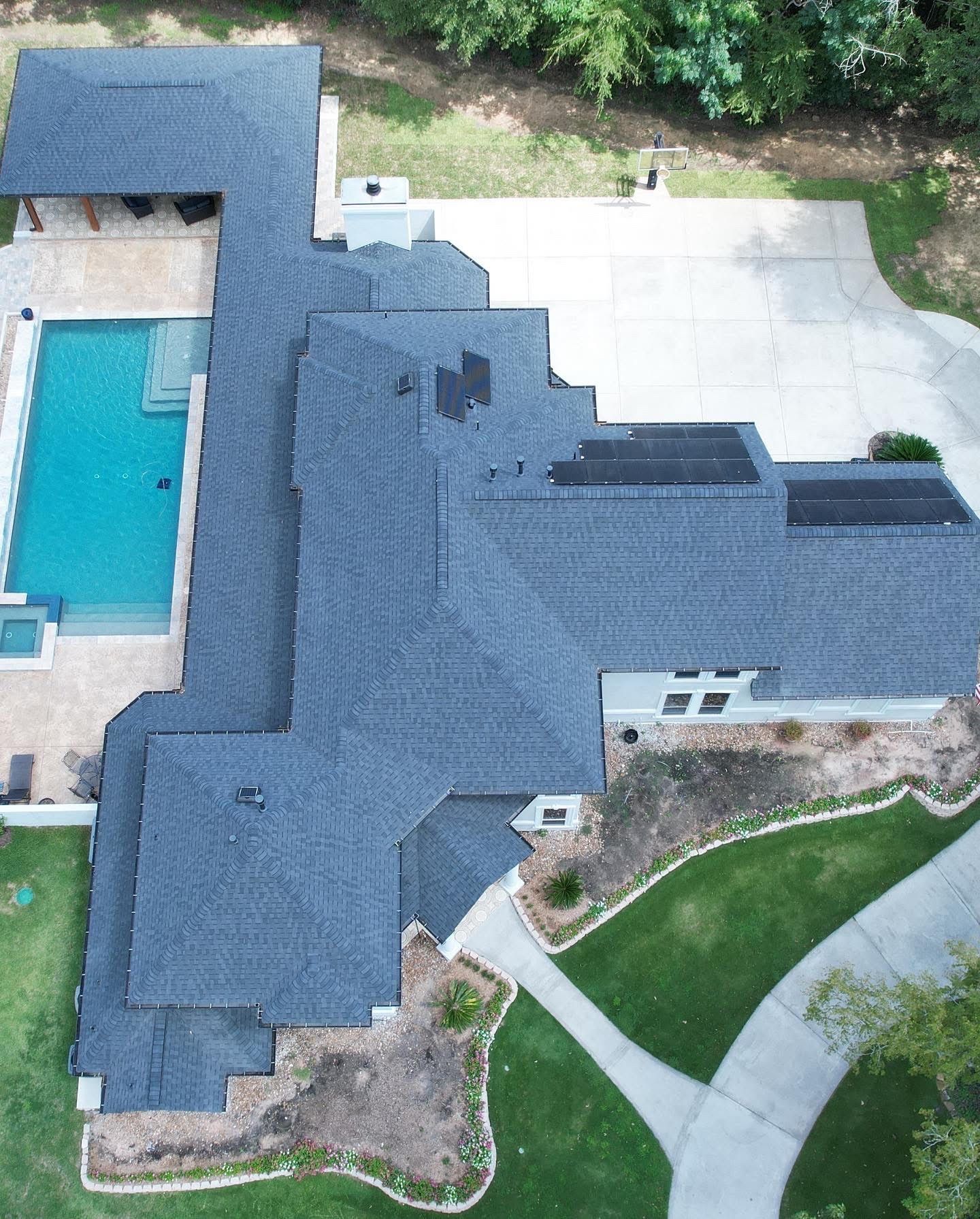 Aerial view of a large house with a complex, dark gray roof, a blue swimming pool, and a concrete driveway.