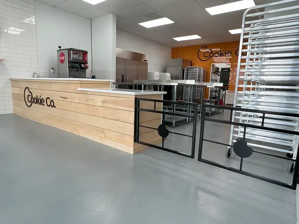 Interior of a Cookie Co. bakery with a light wood counter, beverage dispenser, and industrial kitchen.