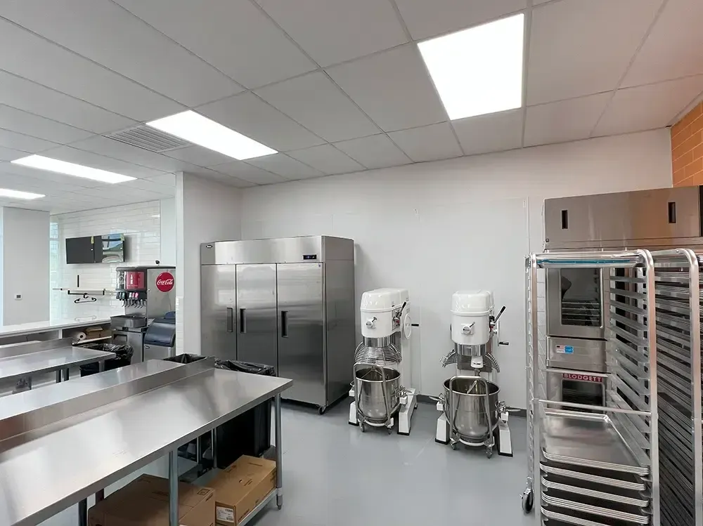 Commercial kitchen with stainless steel equipment. Mixers, refrigerator, work tables, and rolling racks are present.