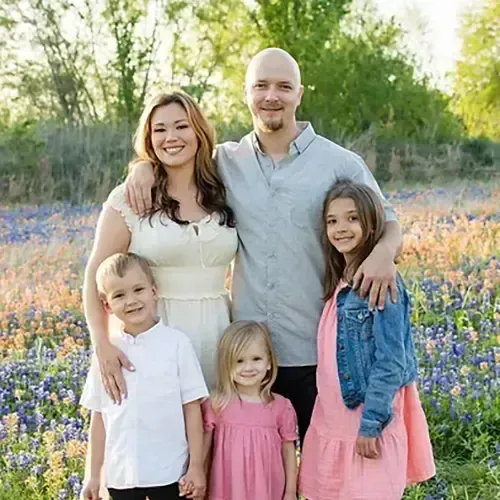 Family posing in a field of wildflowers. Woman in cream dress, man in gray shirt, and three children smile.