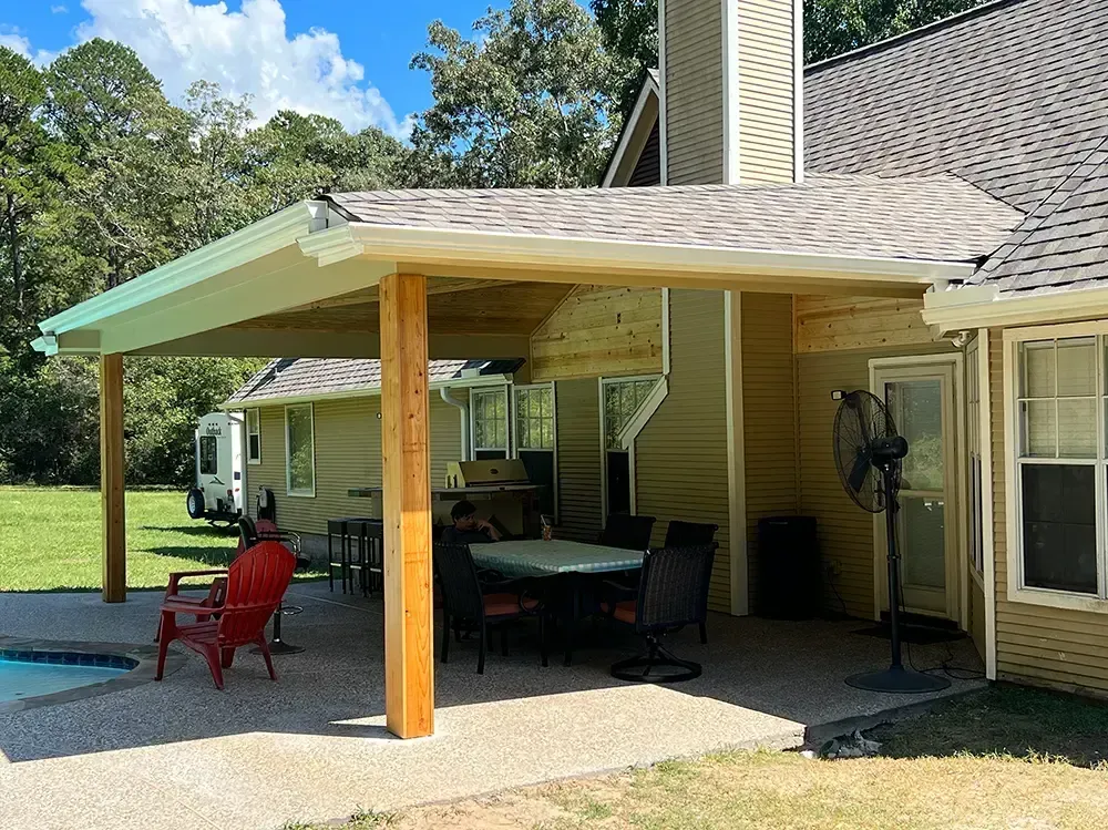 Patio with covered roof, wood supports, and outdoor furniture next to a house on a sunny day.
