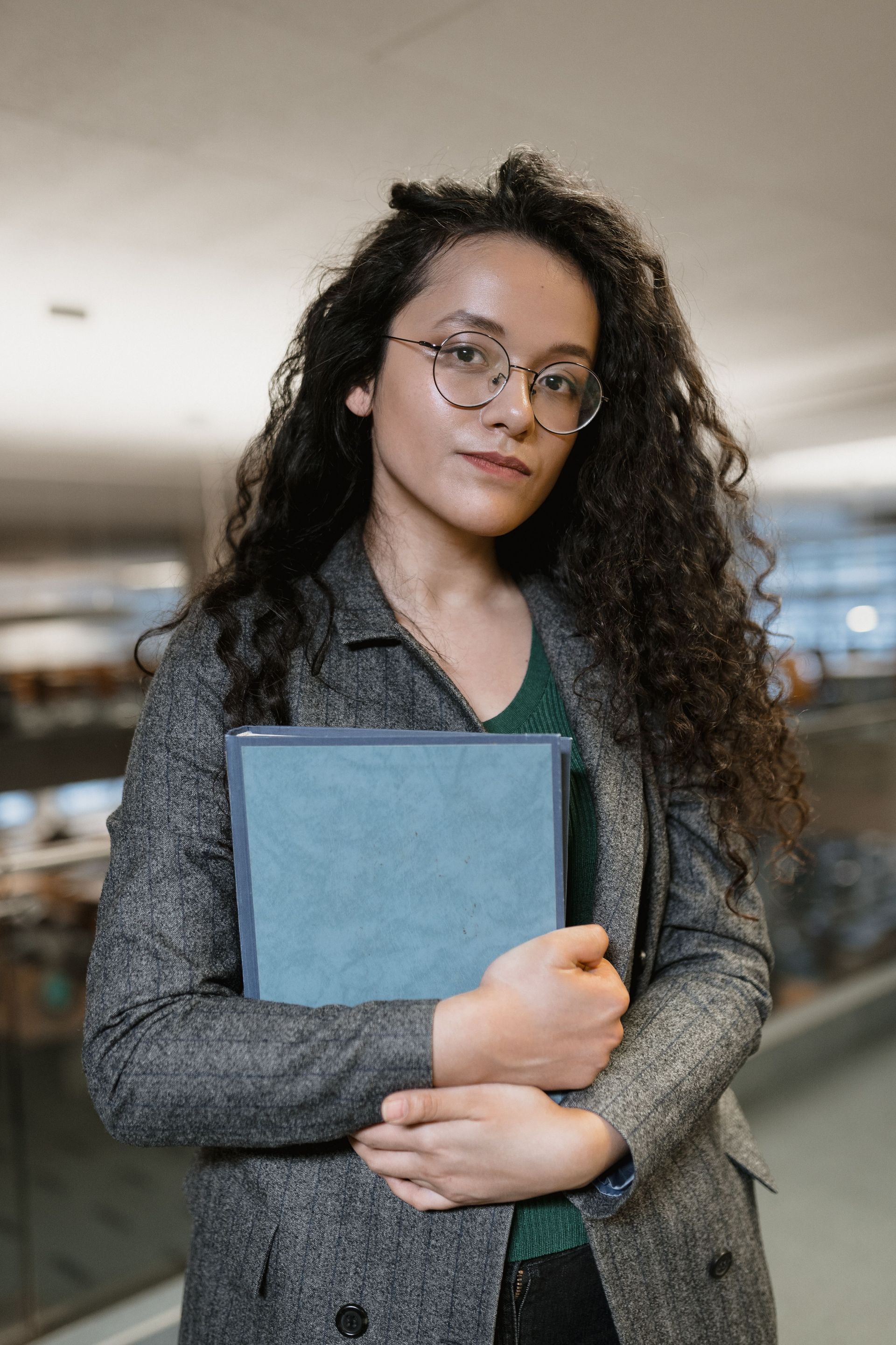 Person in glasses holds a blue folder in a blurred indoor hallway.