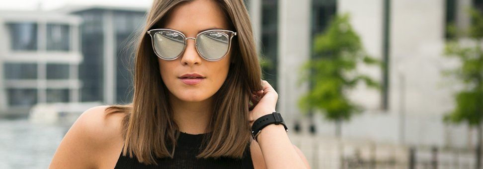 Woman in sunglasses and black top standing outdoors with blurred urban buildings behind her