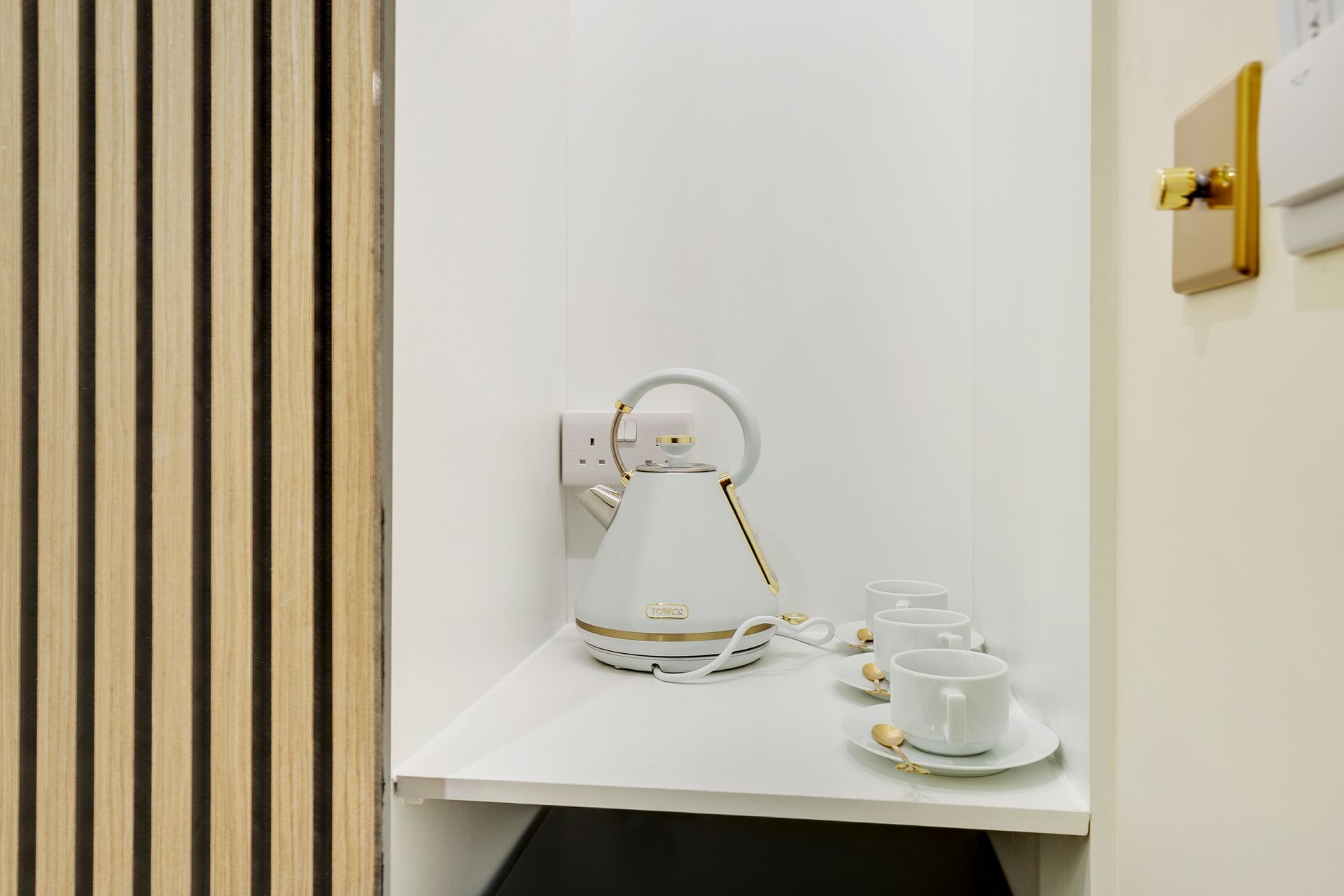 A white kettle and two cups on a shelf in a room.