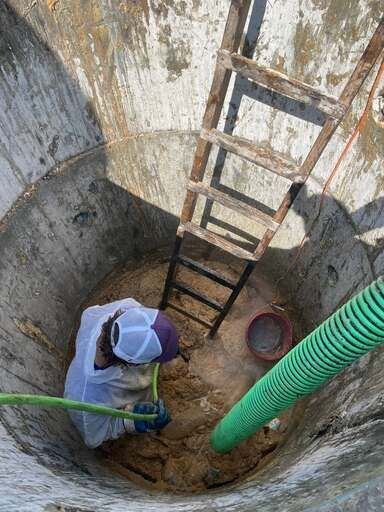 Worker in a concrete well, climbing a ladder beside a green hose and bucket.