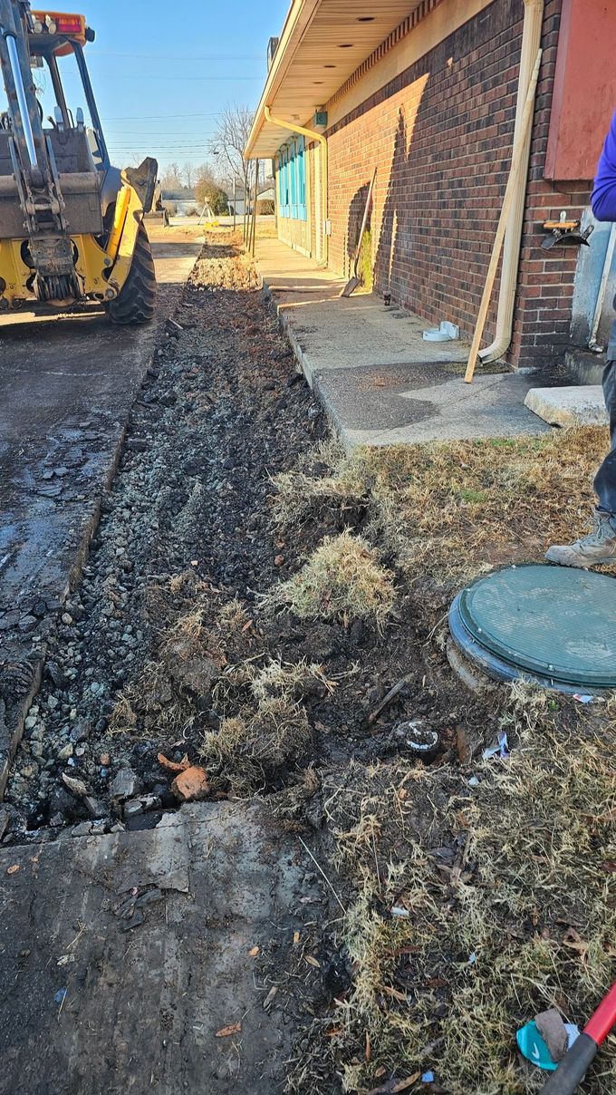 Construction site beside brick building with excavator, muddy trench, and exposed utility covers