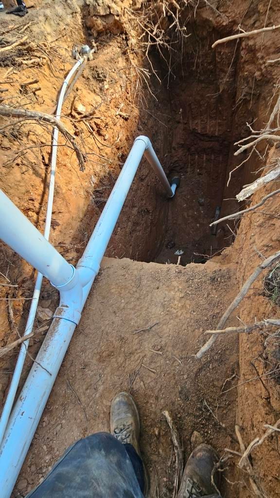 Person standing at the edge of a deep dirt trench with blue pipes running through it