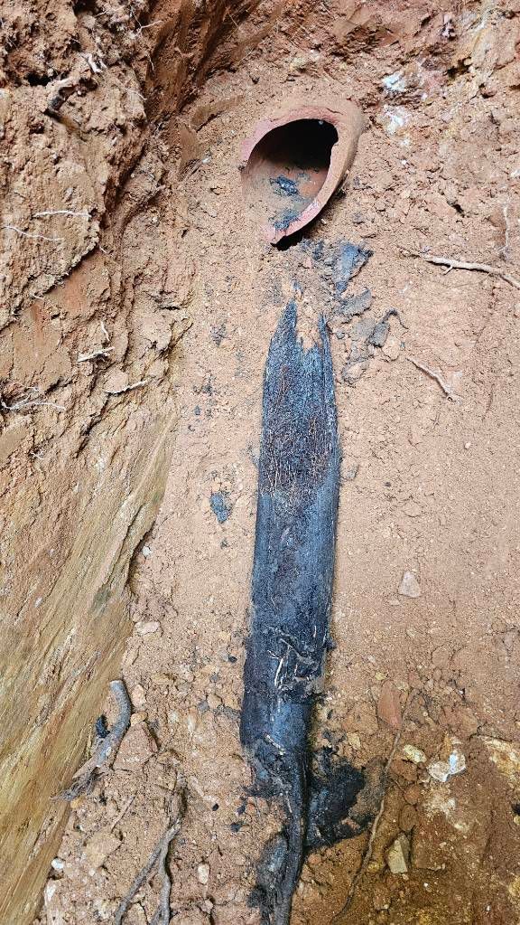 Dark pipe or cable emerging from a round hole in a dirt wall, with a black vertical stain below.