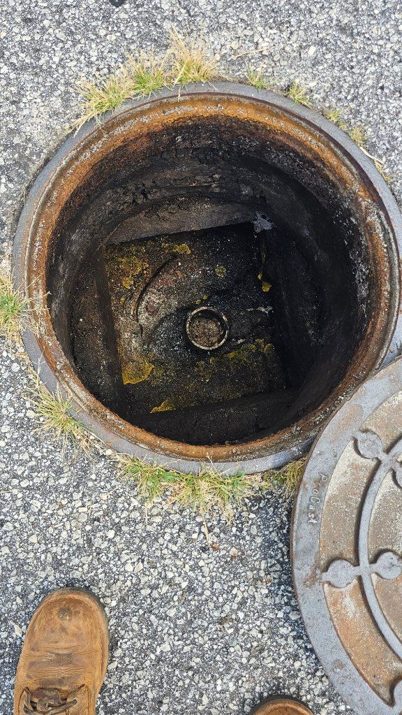 Open manhole or utility access hole in rough pavement, with a boot visible at the lower left.