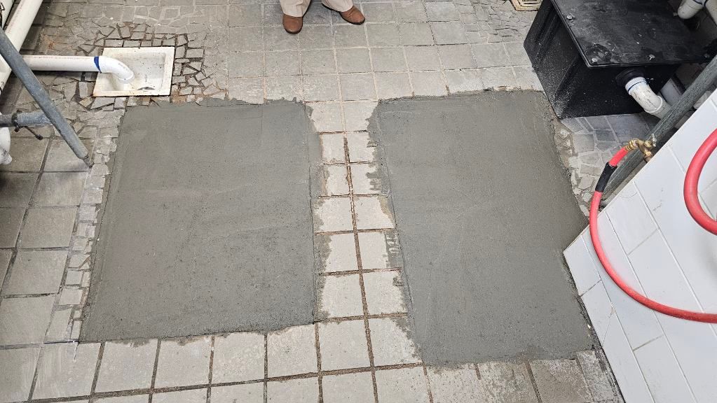 Two gray concrete patches on a tiled walkway beside a white wall and red bucket