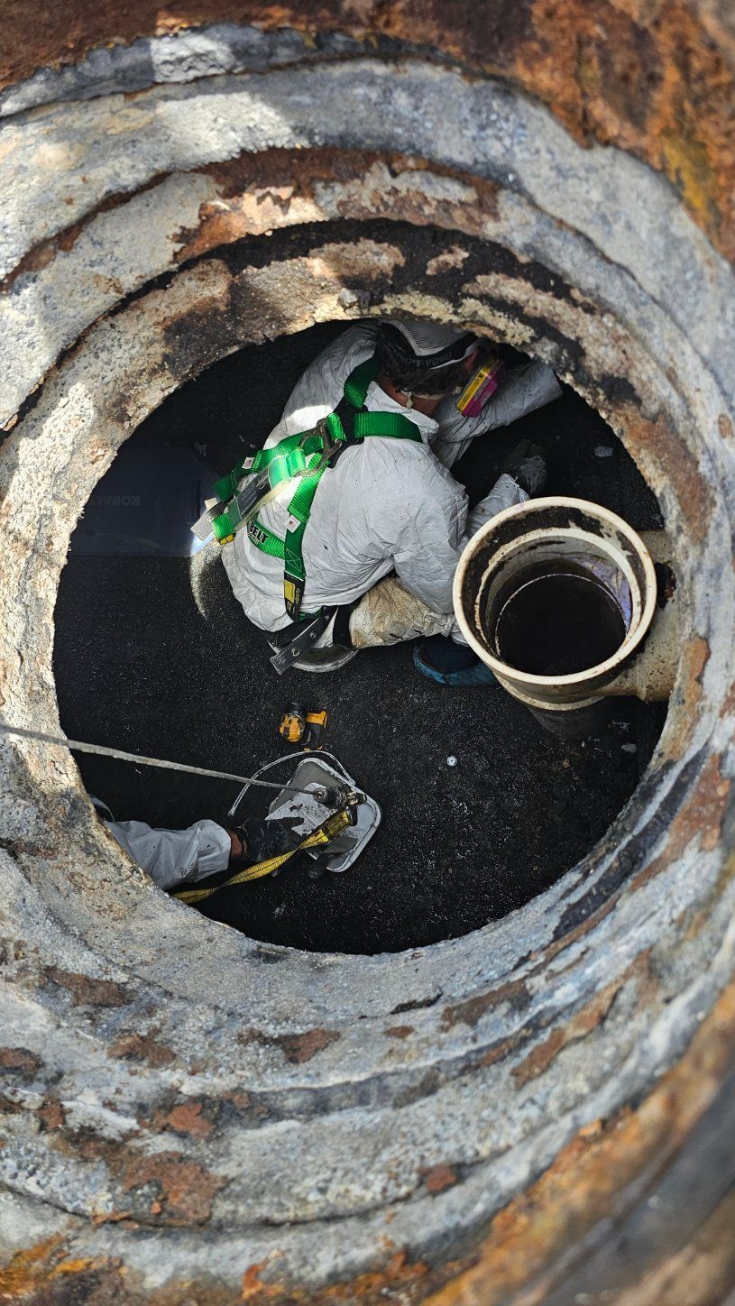 Worker inside a concrete manhole with pipes and cables, viewed from above.