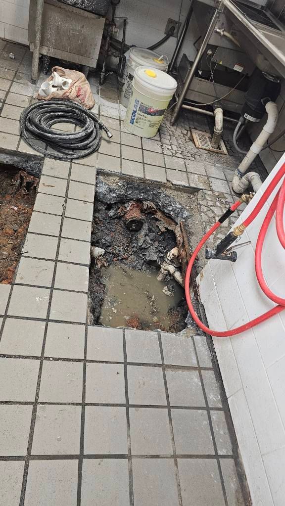 Partially broken tiled floor exposing a large hole in a utility area with tools and hoses nearby