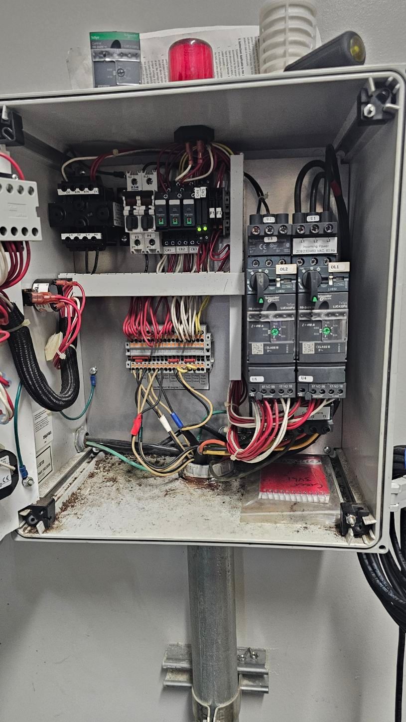 Open electrical control panel with wiring, circuit breakers, and dusty components mounted on a metal post.