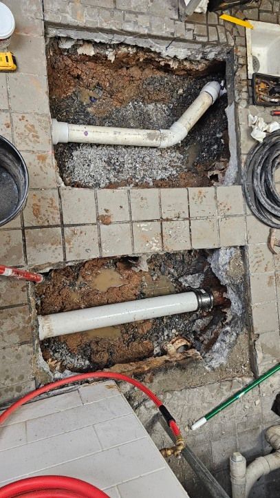 Broken floor tiles exposing white plumbing pipes and soil beneath.