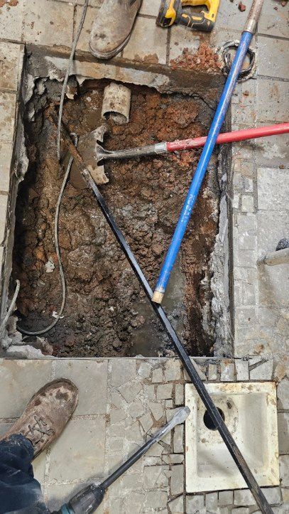 Open trench in concrete floor with exposed soil and plumbing tools, including red and blue pipes.