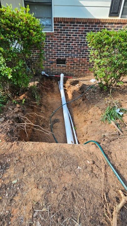 Outdoor trench beside a brick house with a white pipe running through bare dirt and shrubs