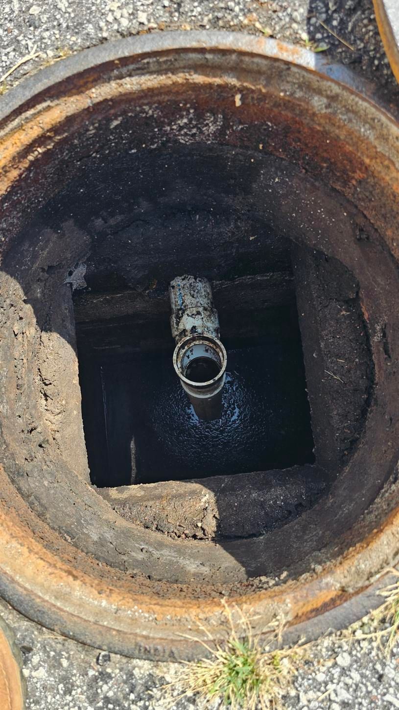 Open rusted manhole showing a dark concrete chamber with a pipe opening in the center.