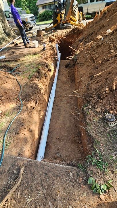 Trench with a white pipe laid in muddy soil at a construction site, workers in the background