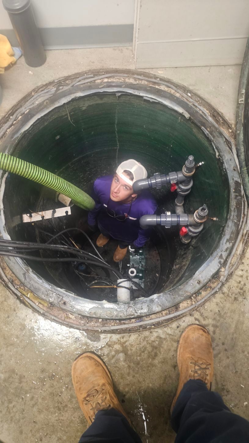 Worker descending into a manhole with hoses and tools for sewer maintenance, viewed from above