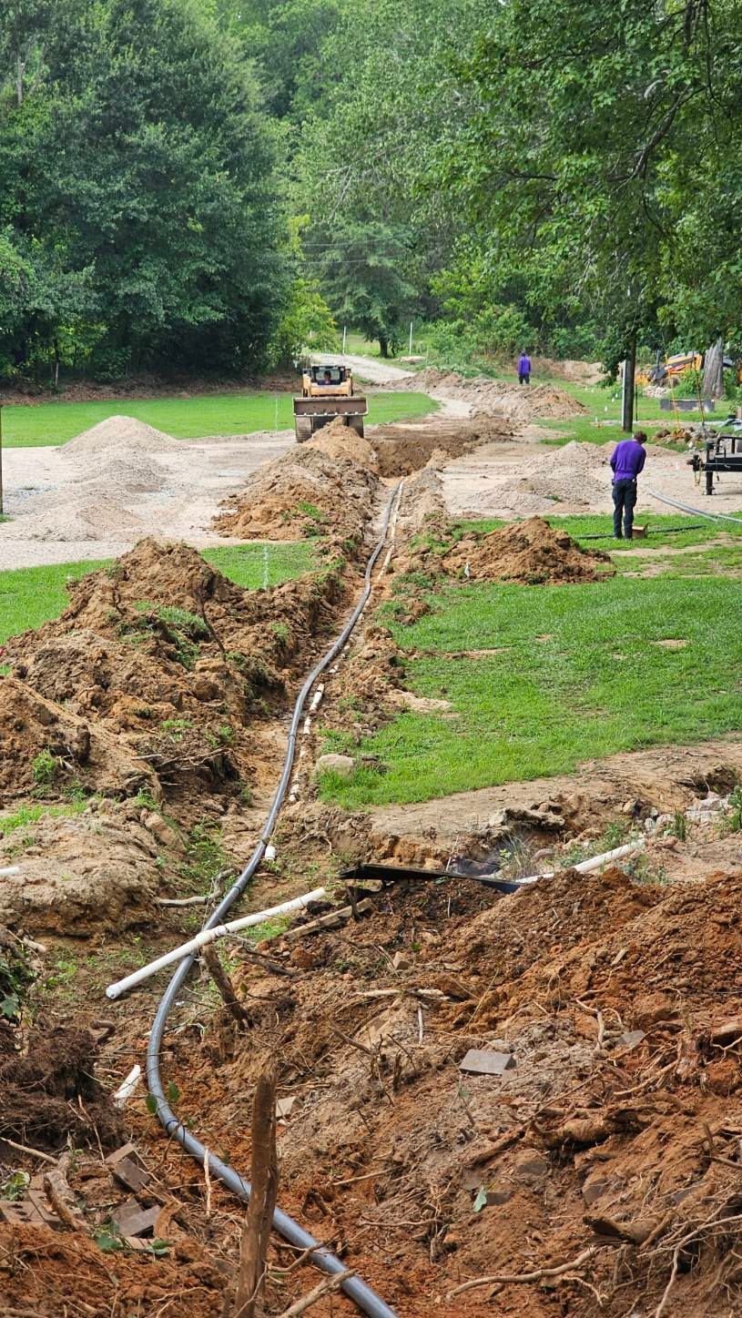 Utility crew trenching a grassy roadside, with pipes laid along the ditch and a backhoe in the distance