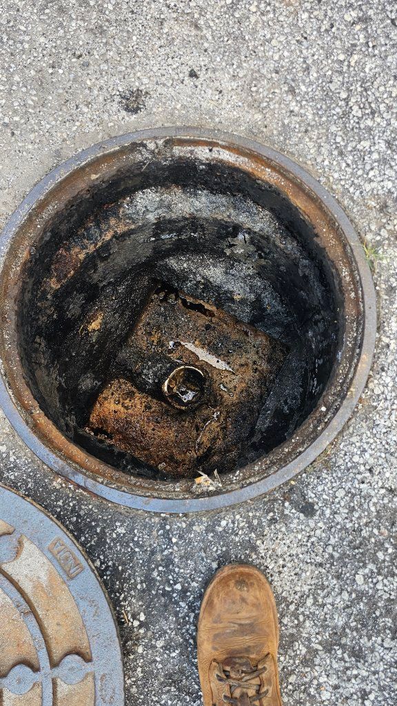 Open manhole with dark, rusted interior on concrete pavement, with a shoe visible at the bottom edge