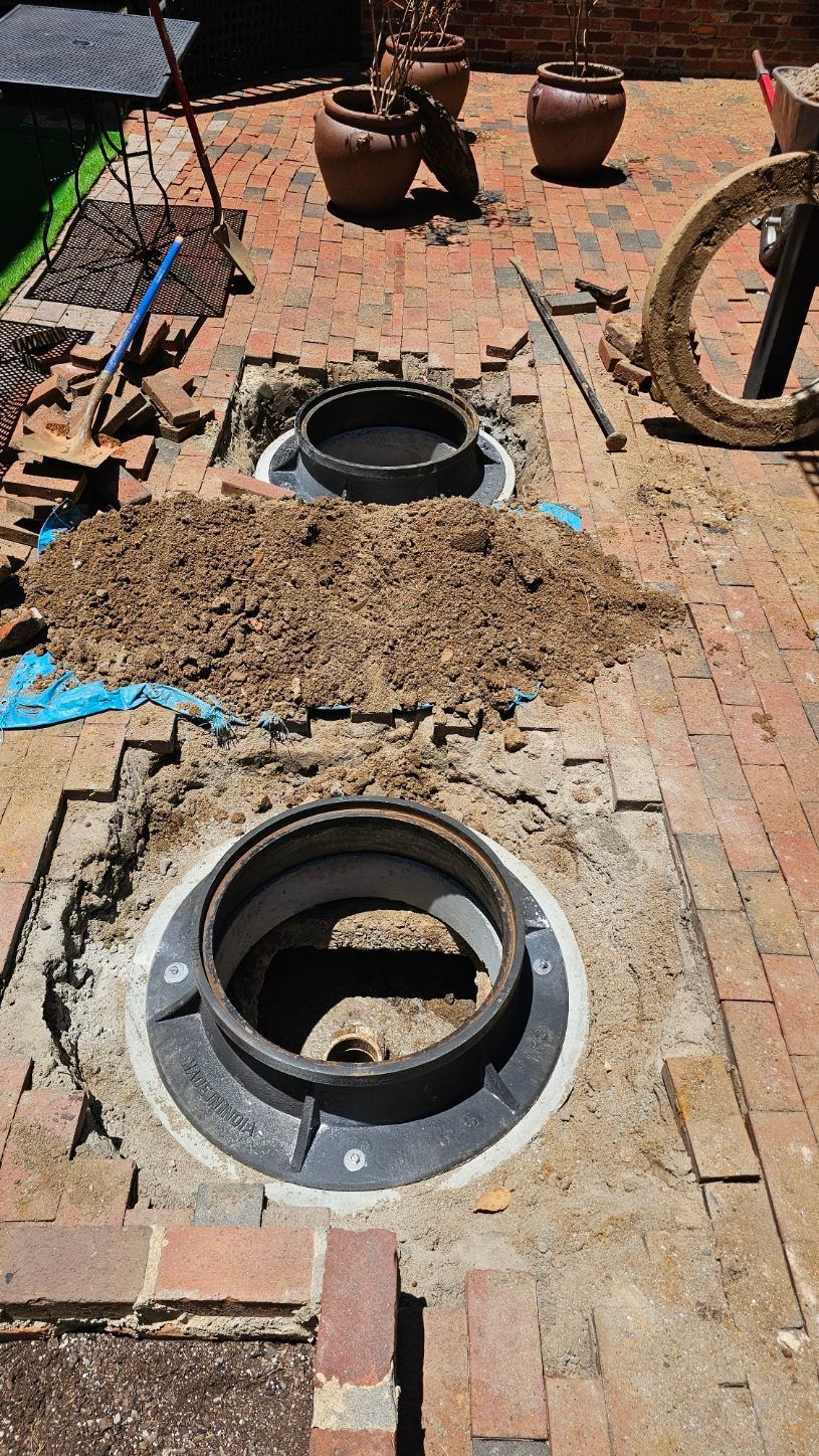 Open manholes in a brick sidewalk beside a dirt pile and potted plants, with construction debris scattered around.