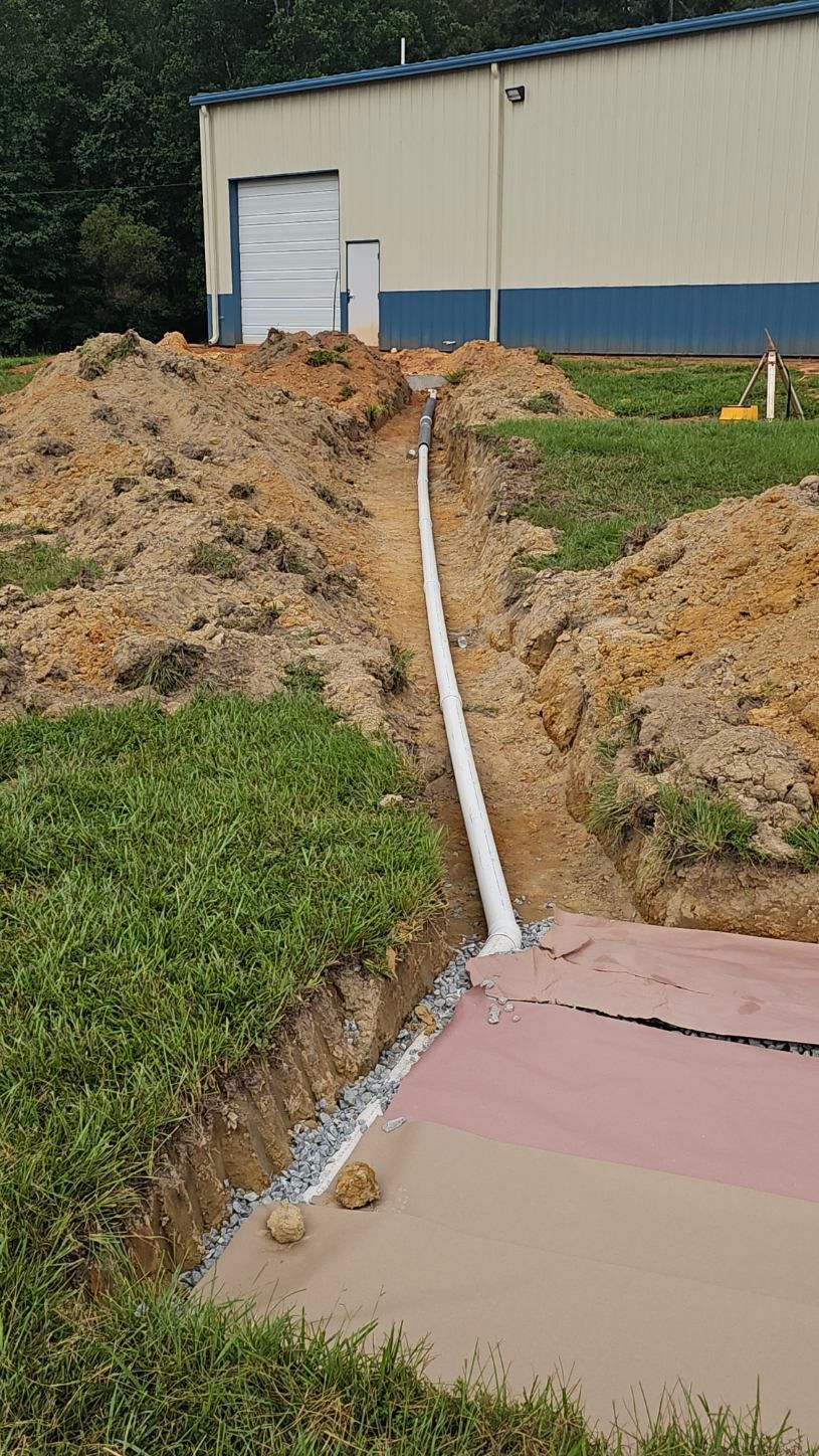 White pipe runs down a dirt slope beside a beige warehouse to a pink and tan sidewalk.