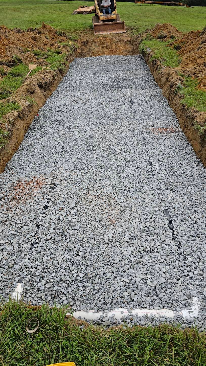 Fresh gravel driveway base in a dug-out trench, with a small excavator in the distance.