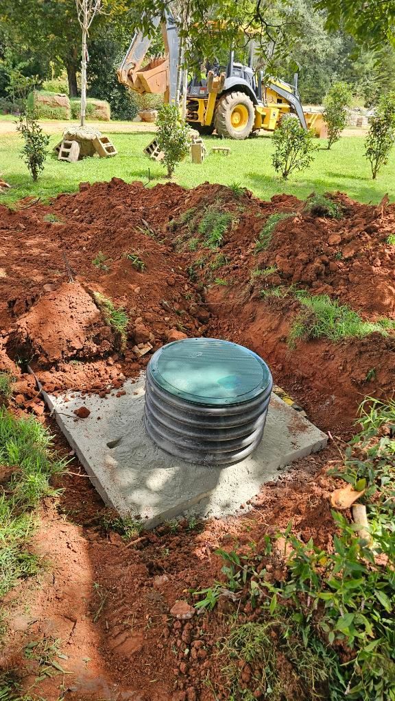 Plastic septic tank lid on a concrete slab amid dug-up soil, with a yellow backhoe in the background.