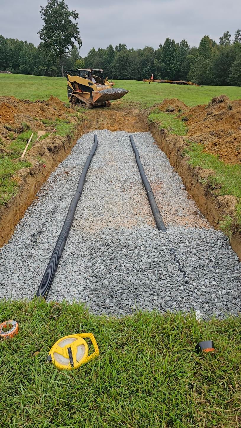 Gravel drainage trench under construction in a grassy field, with a bulldozer in the background.