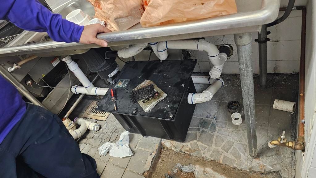 Plumbing under a sink with broken pipes, debris, and a person’s gloved hand holding a tray.