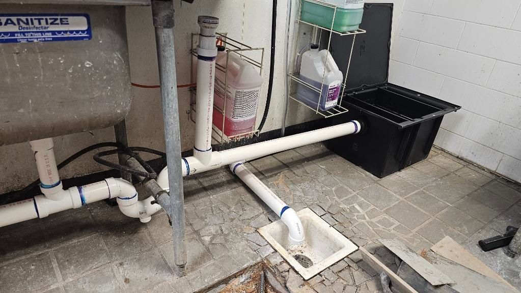 Utility room with exposed white pipes, floor drain, and a black bin against a wall.
