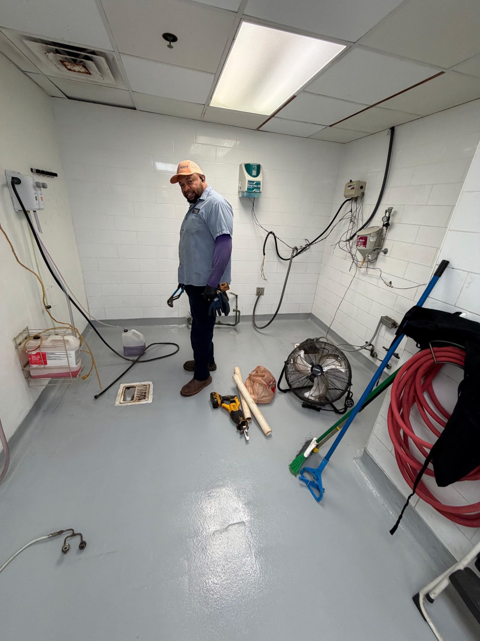 Worker in a utility room with cleaning tools, hoses, and equipment scattered on the floor.