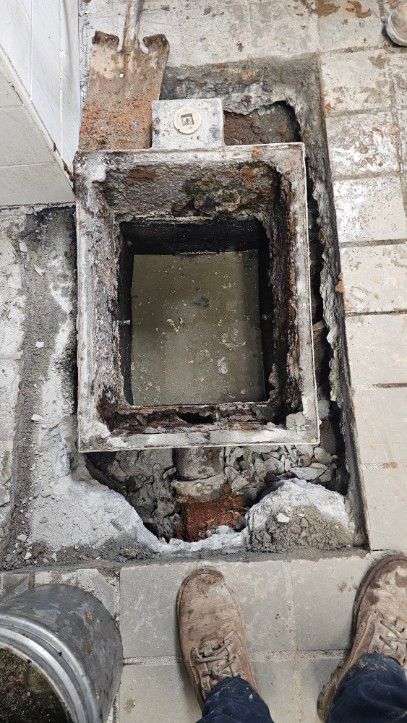 Open concrete floor pit with water, surrounded by debris and tiled pavement, with boots visible at bottom.
