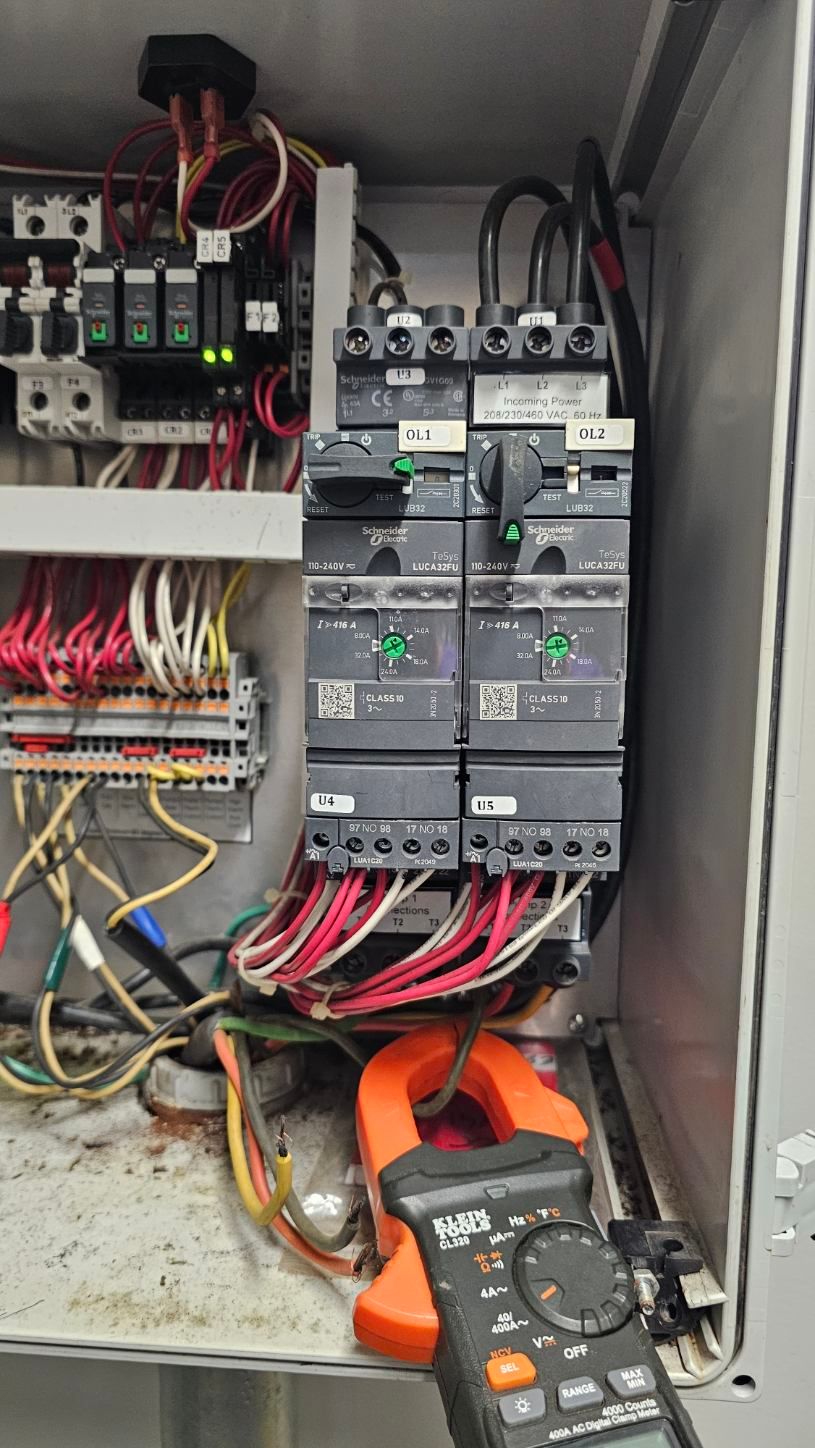 Open electrical control cabinet with wiring, circuit breakers, and a clamp meter at the bottom.