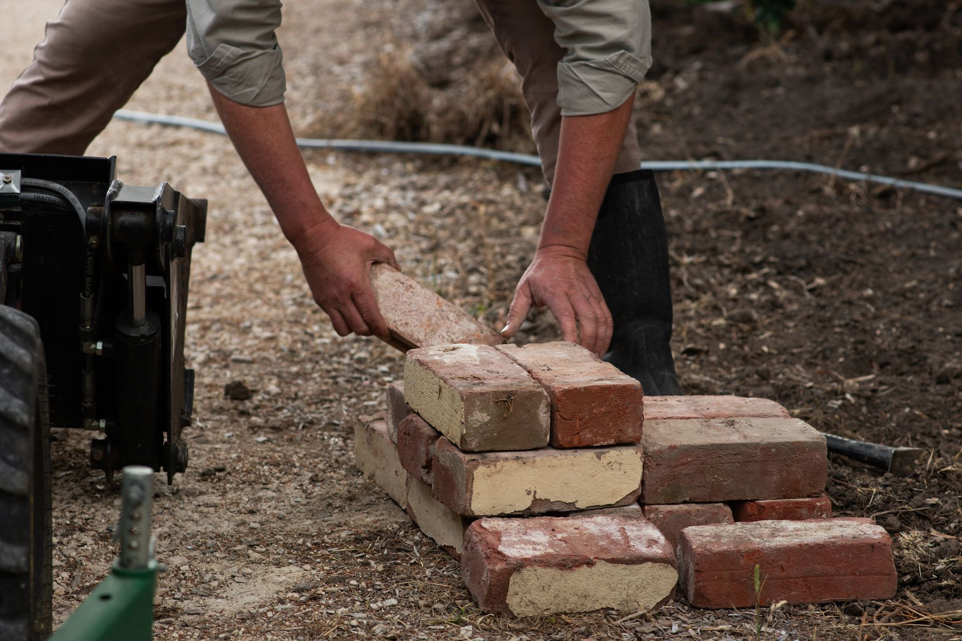 ‘’Person stacking red and beige bricks on a dirt ground’’