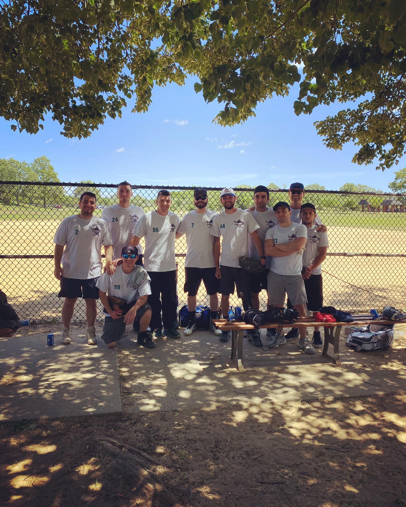 Group posing outdoors under trees beside a chain-link fence, with sports gear and a bench in front
