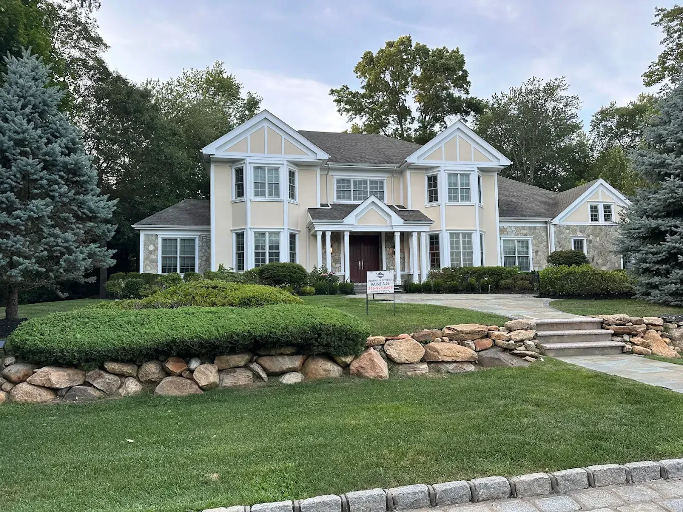 Large beige house with white trim, stone retaining wall, and manicured front lawn in a suburban neighborhood