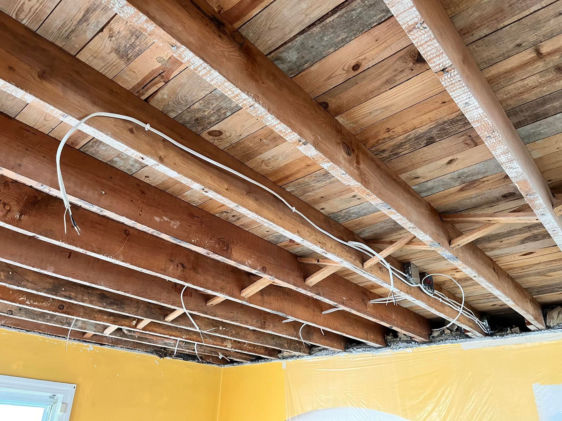 Exposed wooden ceiling beams with electrical wiring above yellow walls in an unfinished room