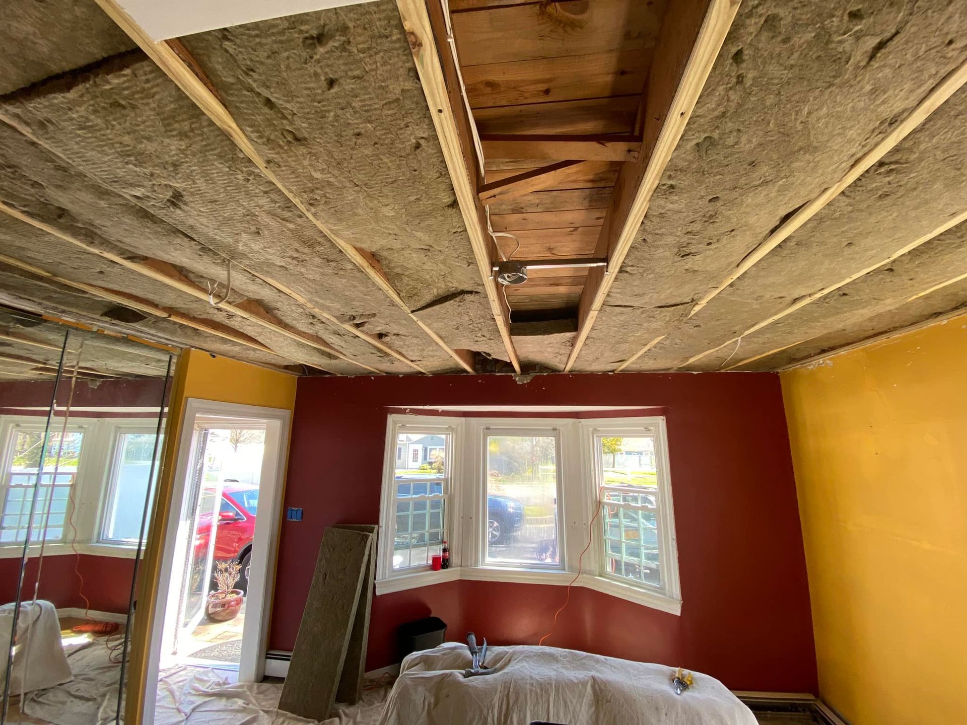 Interior room under renovation with exposed ceiling insulation, red and yellow walls, and a windowed bay area.