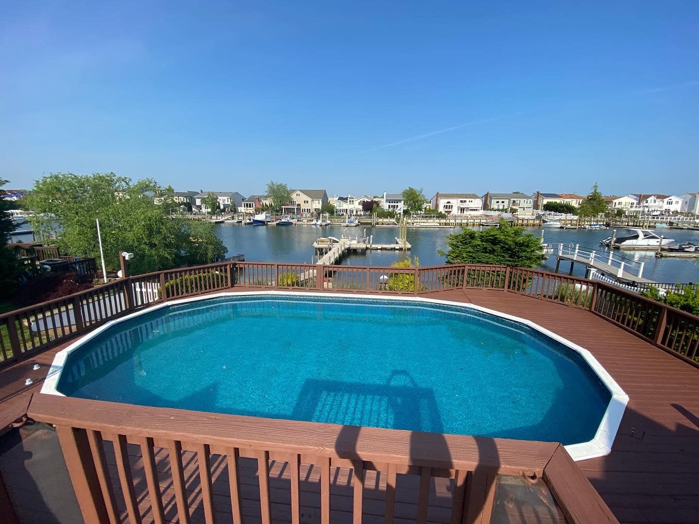 Backyard deck with oval pool overlooking a calm waterfront and houses under a clear blue sky