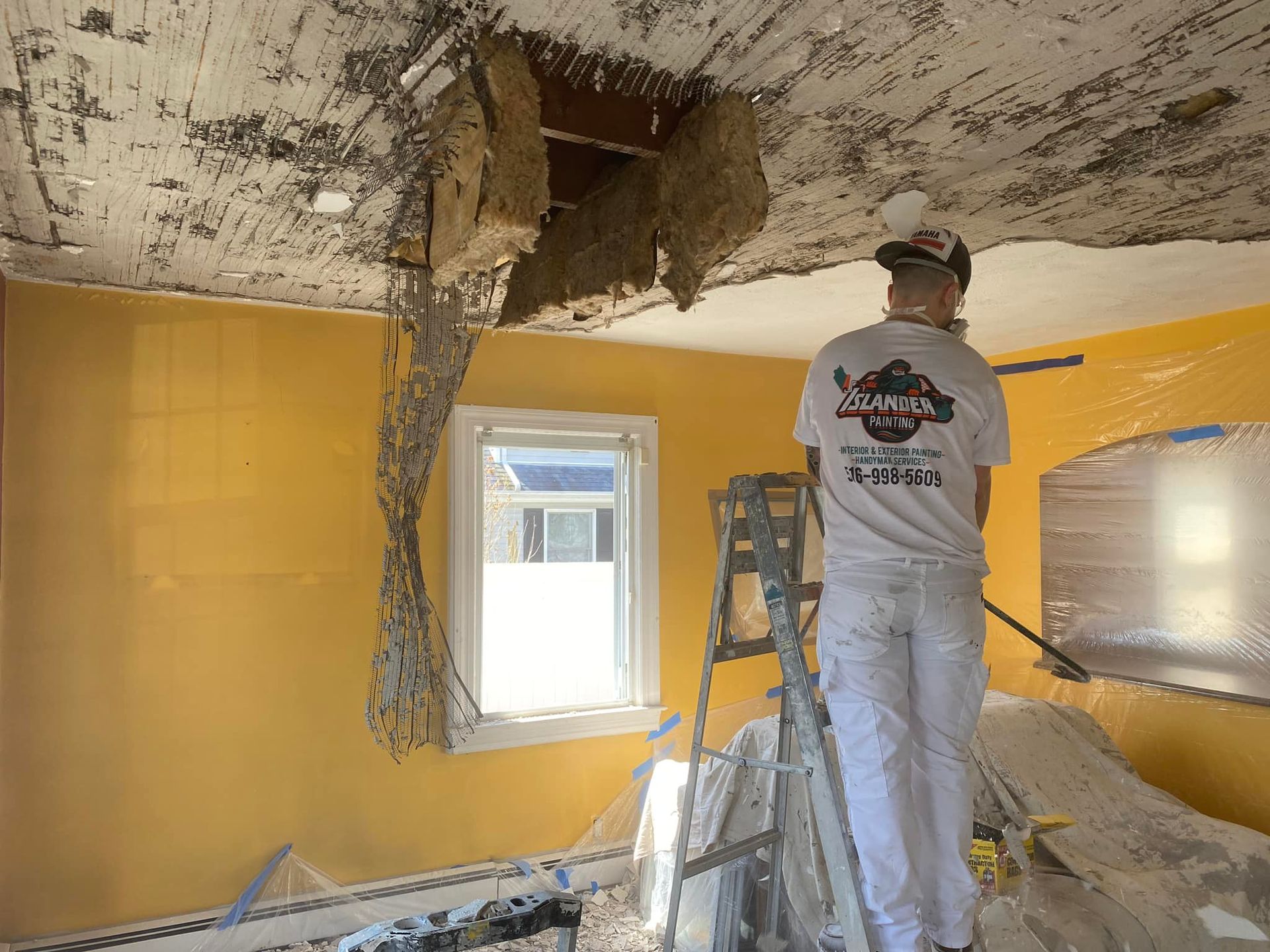 Worker on ladder removing damaged ceiling in yellow room with debris on floor.