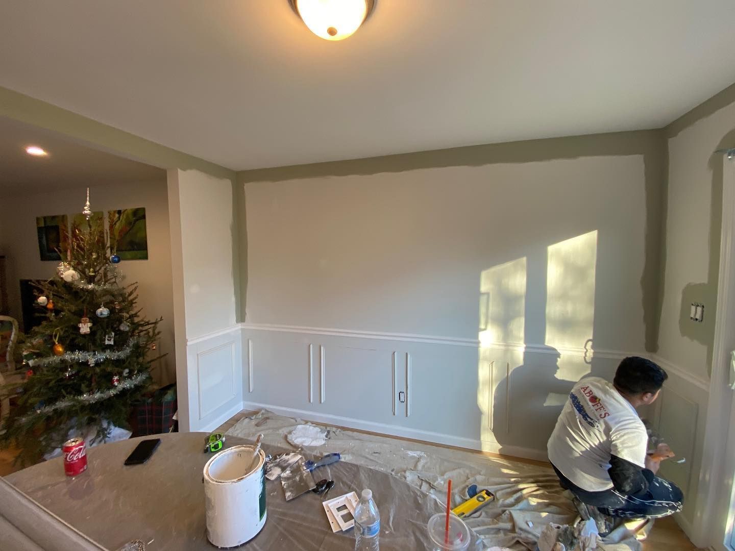 Person painting a dining room wall beside a Christmas tree, with floor covered in drop cloths and supplies