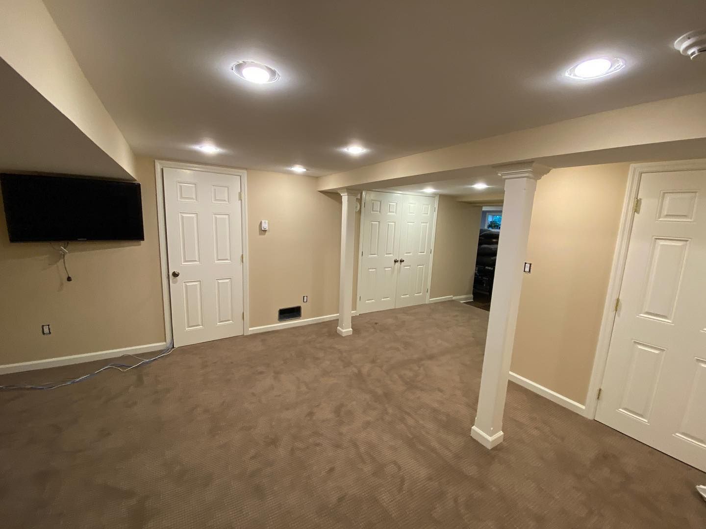 Empty beige basement room with carpet, recessed lights, white doors, and a wall-mounted TV