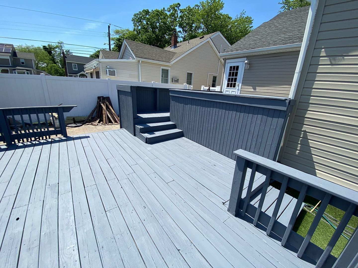 Gray wooden deck with steps and railings, overlooking a yard and neighboring houses on a sunny day