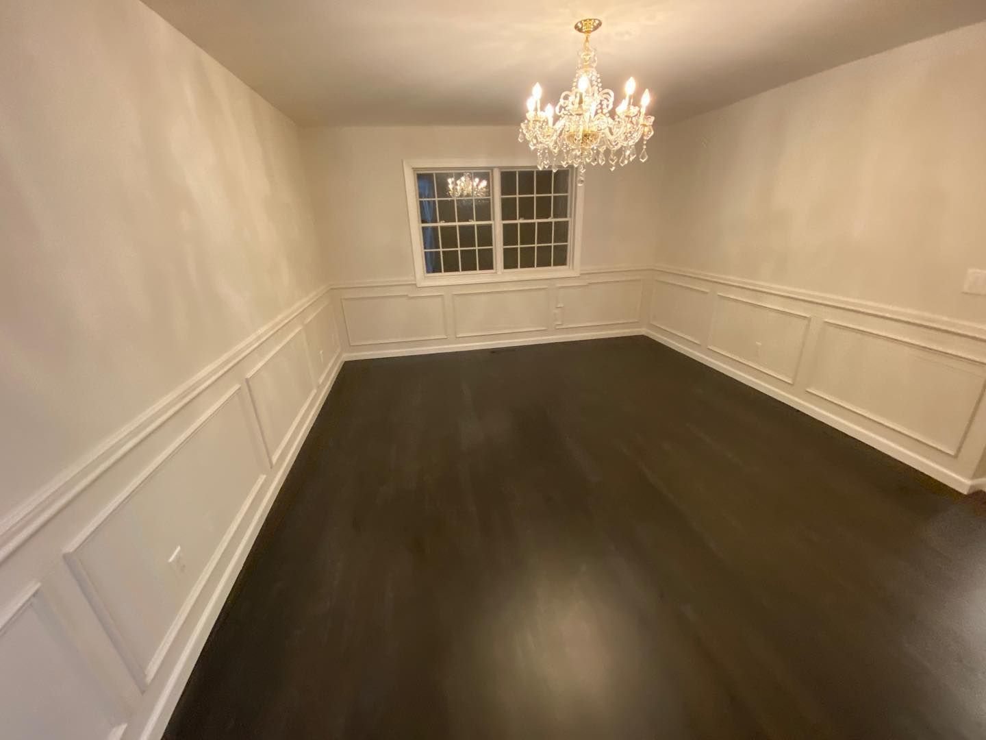 Empty dining room with dark hardwood floors, white wainscoting, chandelier, and a window at the far end